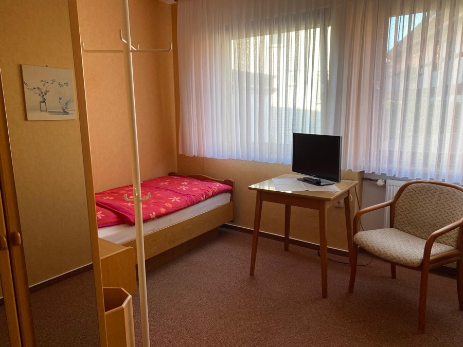 Einzelzimmer mit Fernseher, Sessel und Gardine